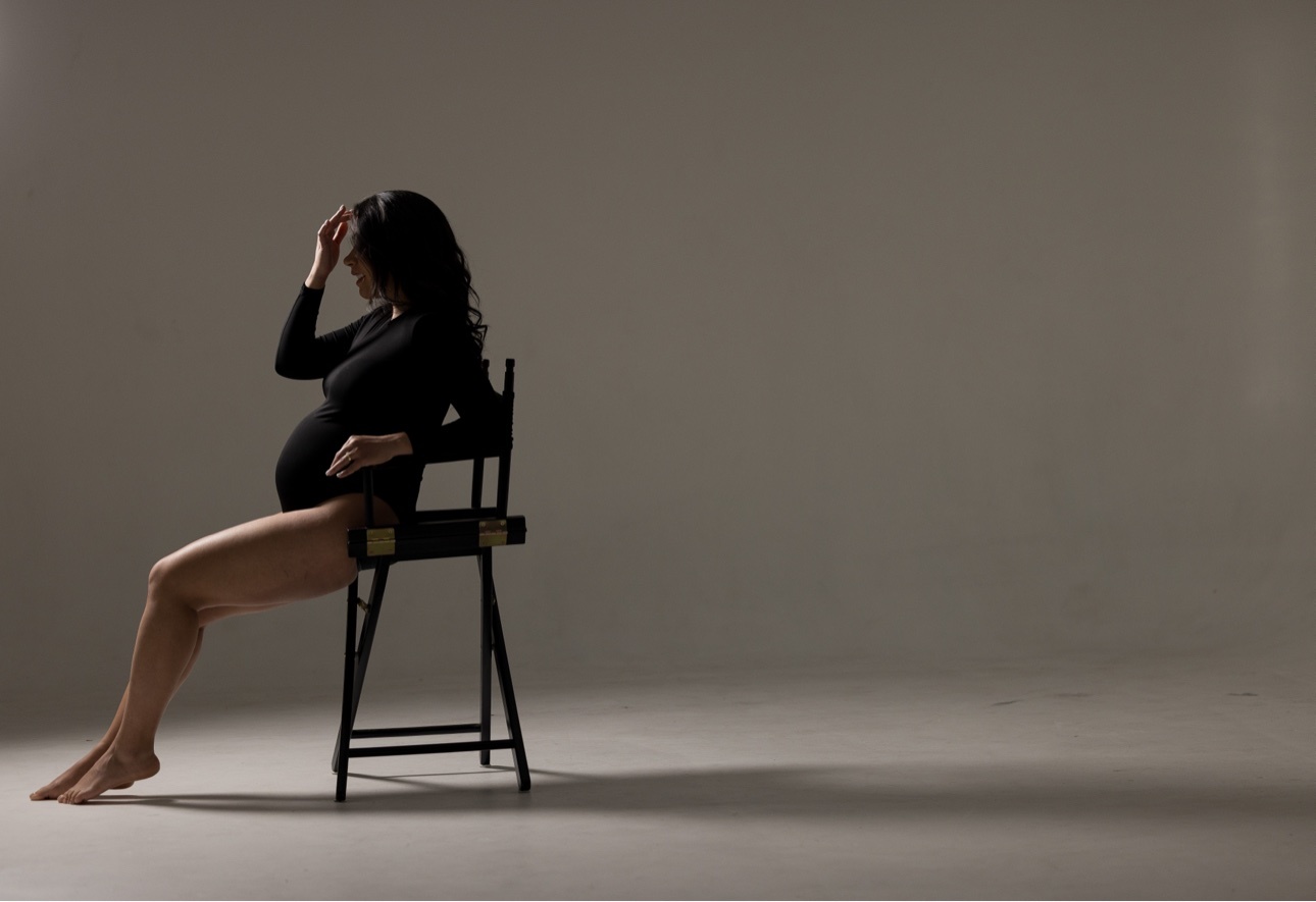 Color studio portrait of an expectant mother in a black bodysuit seated in profile on a director's chair, her whole body and bare feet fully visible with floor beneath them against an empty warm taupe backdrop