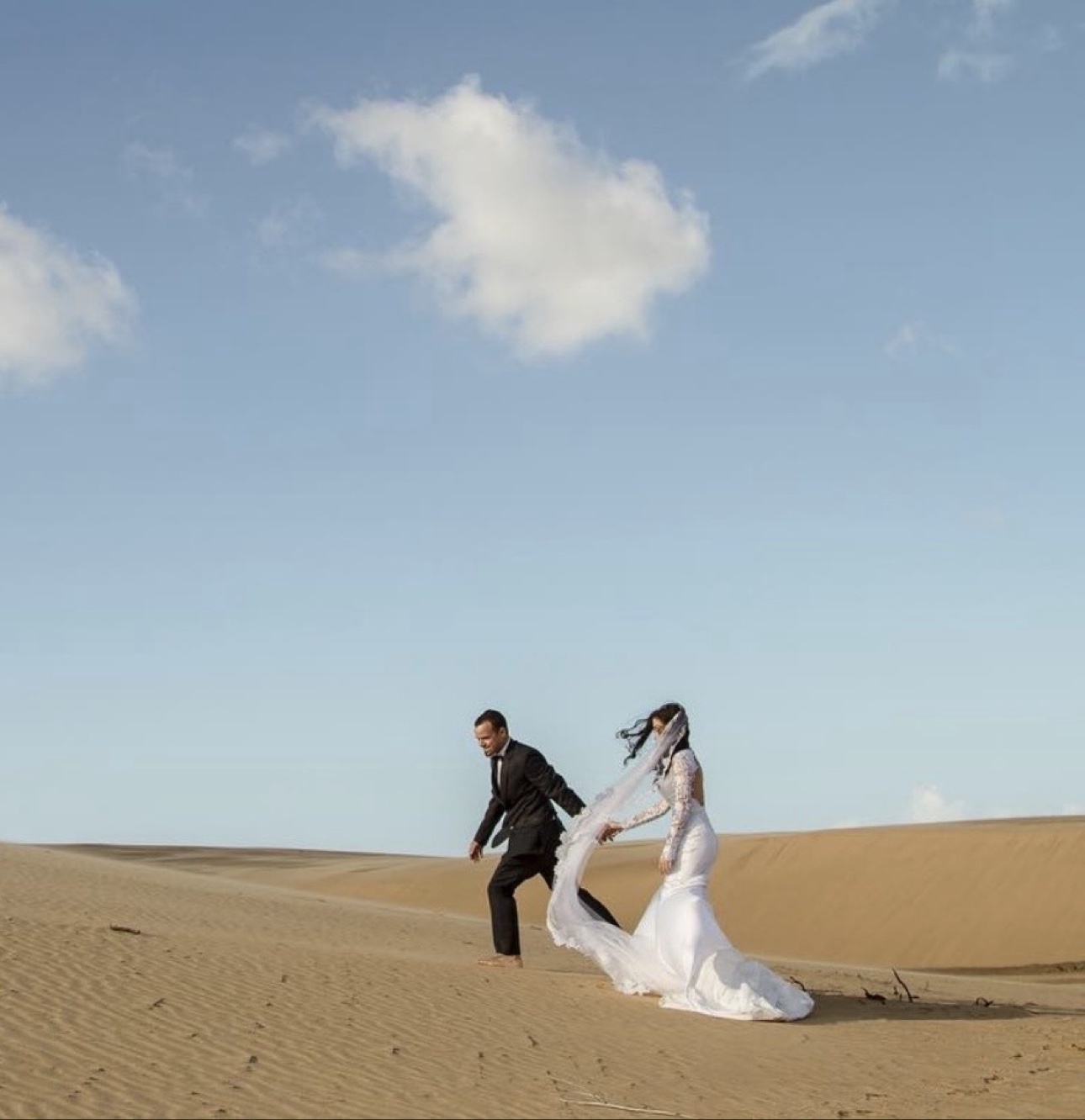 Newlyweds walking barefoot together across sweeping sand dunes