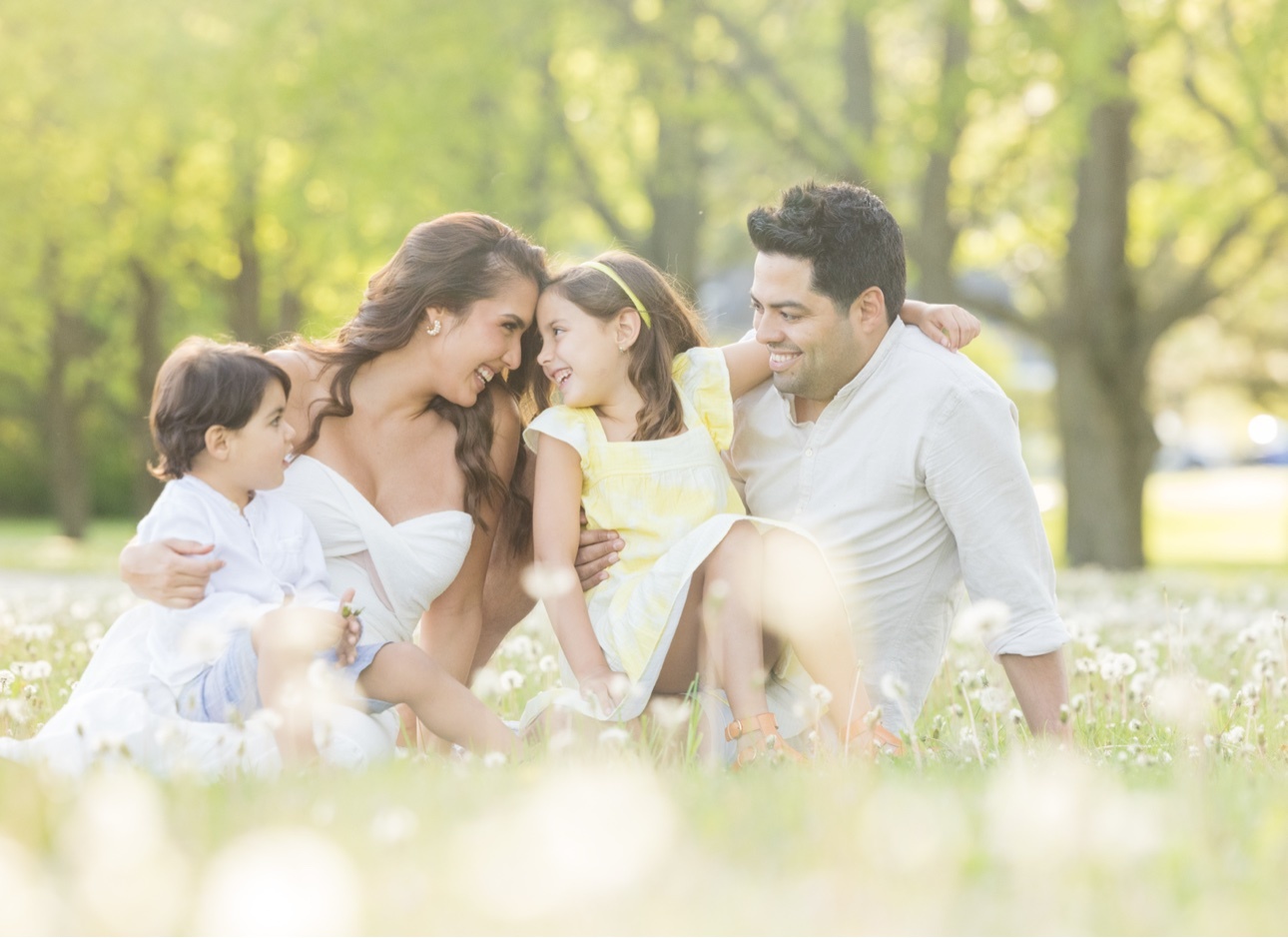 Family of four — mother, father, their daughter in a yellow dress, and their young son — laughing together in a sunlit field of dandelions