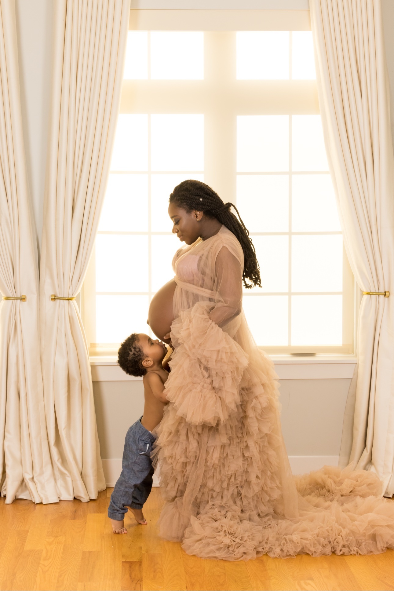 Expectant mother in a beige tulle gown standing before a bright window as her young son kisses her belly